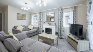 Ein Wohnzimmer mit grauem Sofa, Couchtisch, Kamin, Fernseher auf Holzständer und floralem Vorhang.