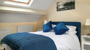 Un dormitorio con tragaluz, una cama con ropa de cama azul y blanca, una mesita de noche con lámpara y un cuadro enmarcado en la pared.