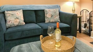 Una sala de estar con un sofá azul, cojines estampados, una bandeja de mimbre con una botella y una copa de vino, y una lámpara.
