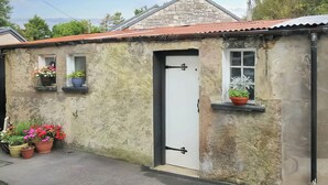 Un pequeño edificio de piedra con techo inclinado, una puerta blanca y ventanas con plantas en macetas.