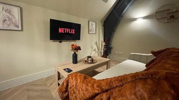 A modern room with a flat-screen TV displaying 'NETFLIX', a wooden bedside table with a vase of flowers, a bed with a brown blanket, and a window with a view of the sky. - Chic Apartment on The Headrow, Leeds (Leeds)