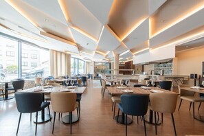 A modern dining area with tables and chairs, large windows, and a kitchen in the background. - Jupiter Porto Hotel - Rooftop & Spa (Porto)