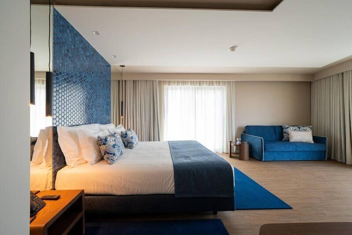 Habitación de hotel moderna con una cama grande, un sofá azul y vistas a la ciudad.