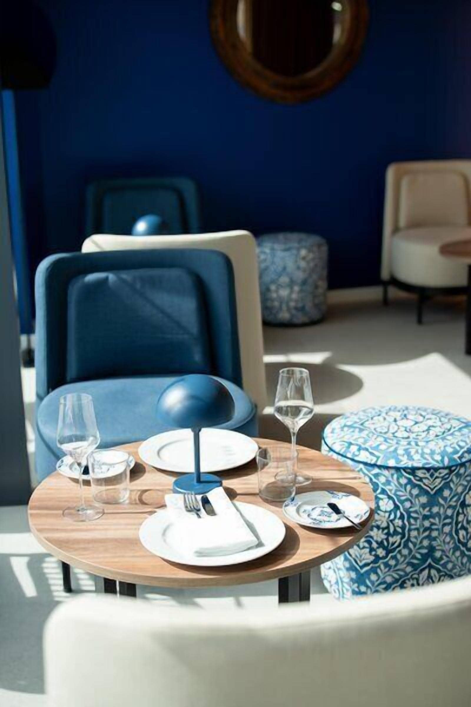 A dining table set with plates, glasses, and cutlery, surrounded by chairs and ottomans in a room with blue walls and a round mirror.