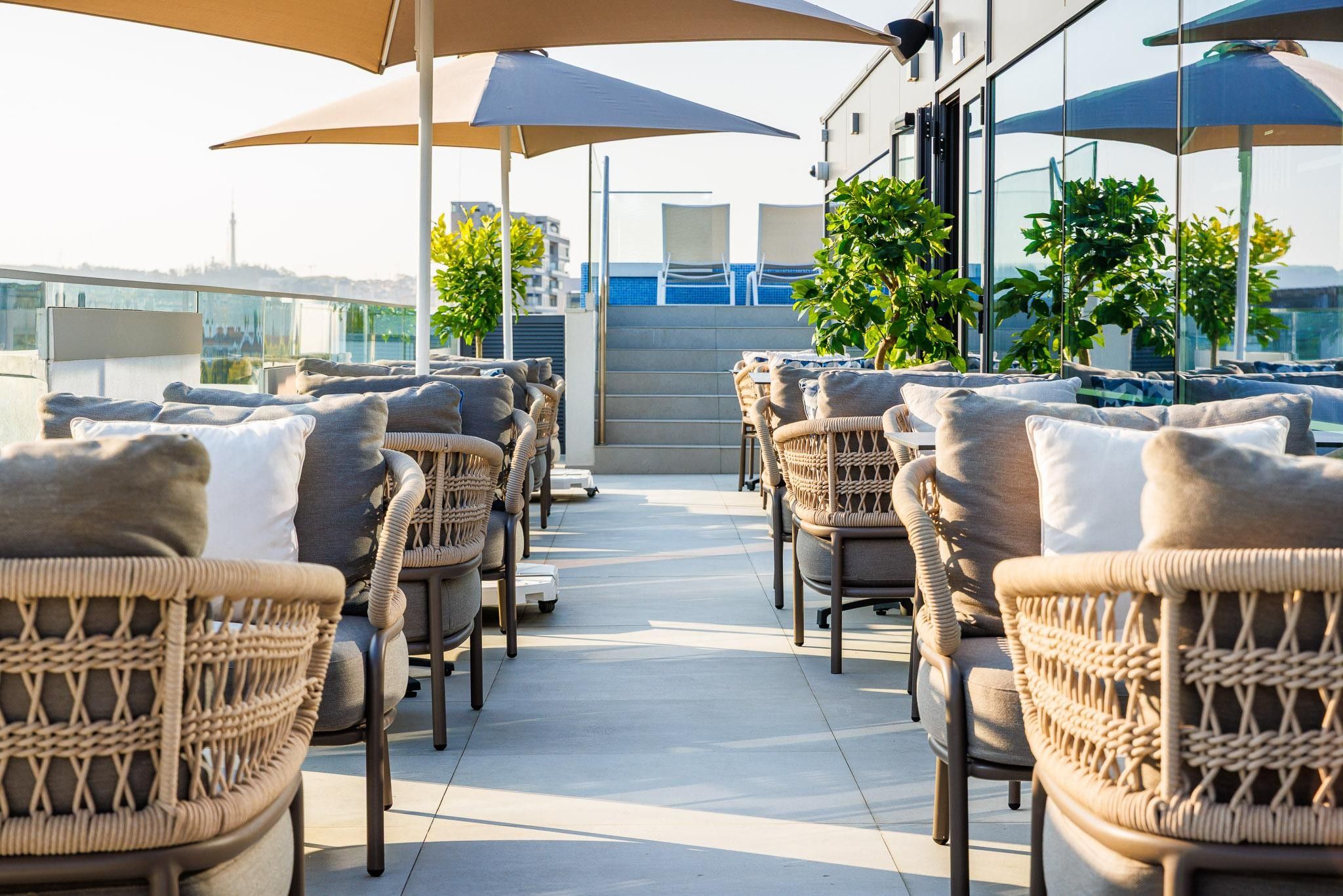 Eine Reihe von Korbstühlen mit Kissen, auf einer gefliesten Terrasse, mit Sonnenschirmen, die Schatten spenden.