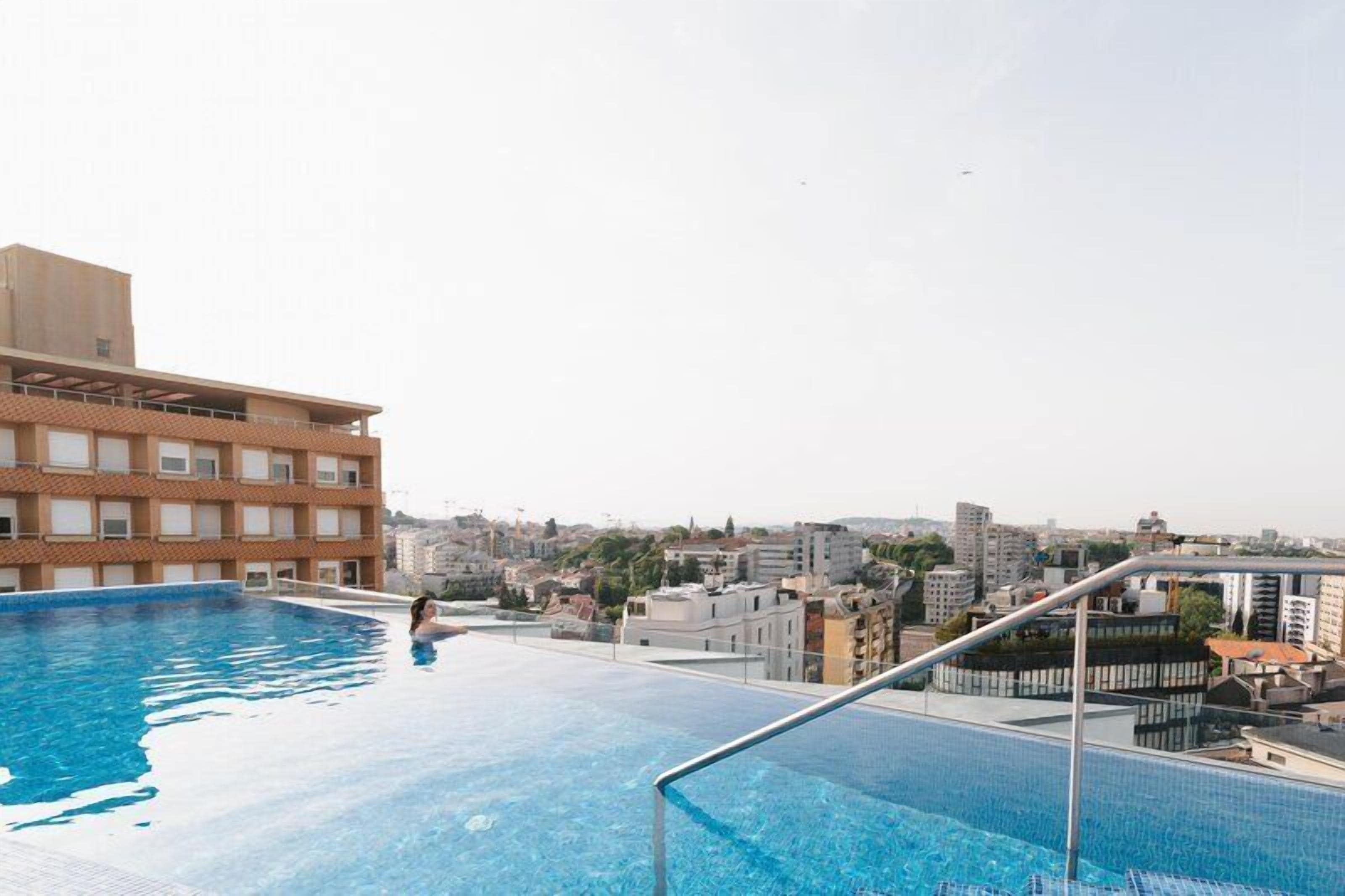Una piscina infinita en la azotea con vistas a la ciudad, una persona nadando y un edificio con balcón.