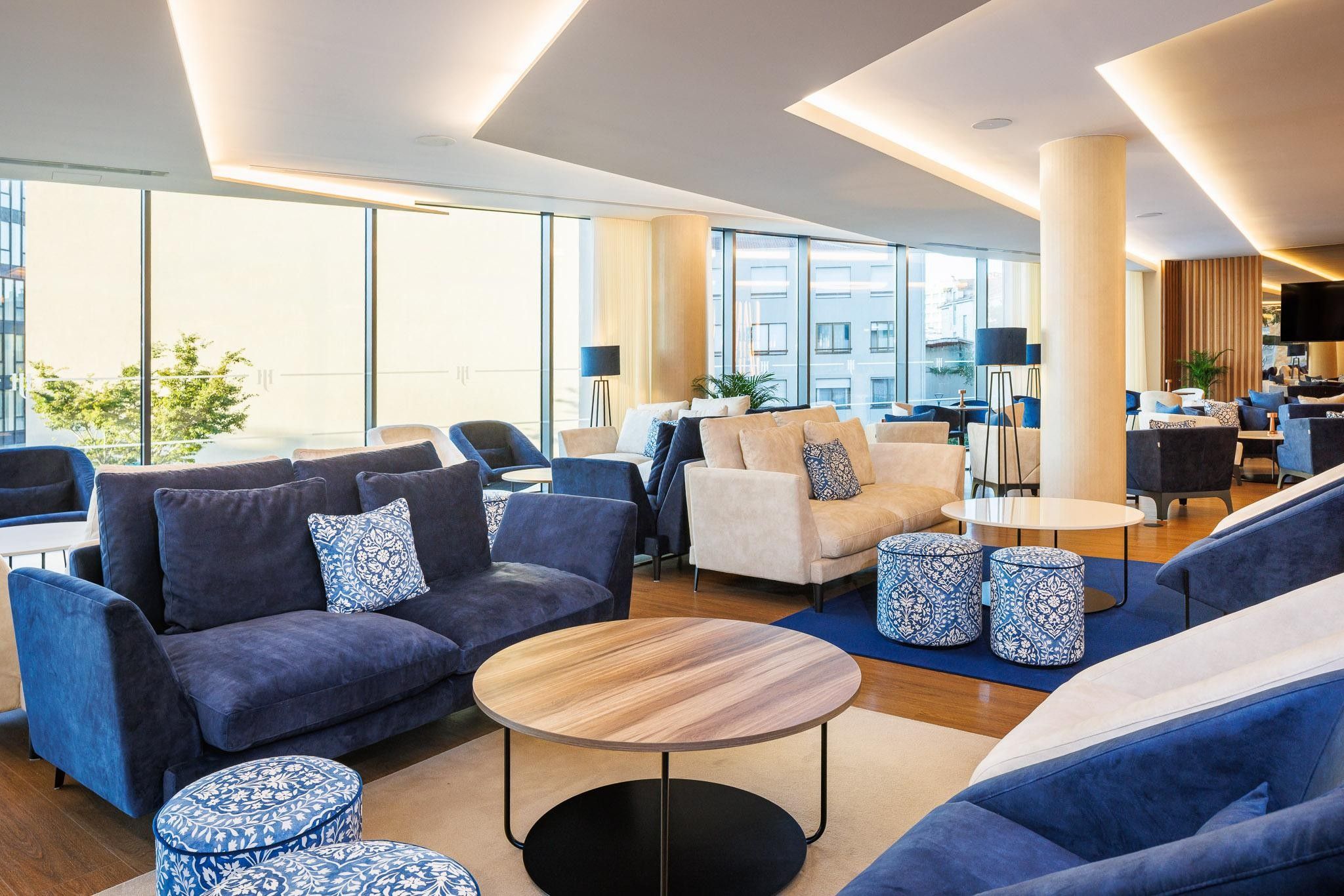 A modern lounge area with sofas, ottomans, and a coffee table, featuring large windows and a city view.