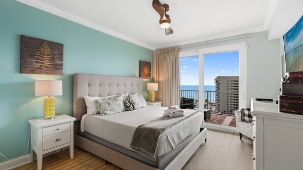 A bedroom with a large bed, two bedside tables, a ceiling fan, and a balcony with a view of the sea.