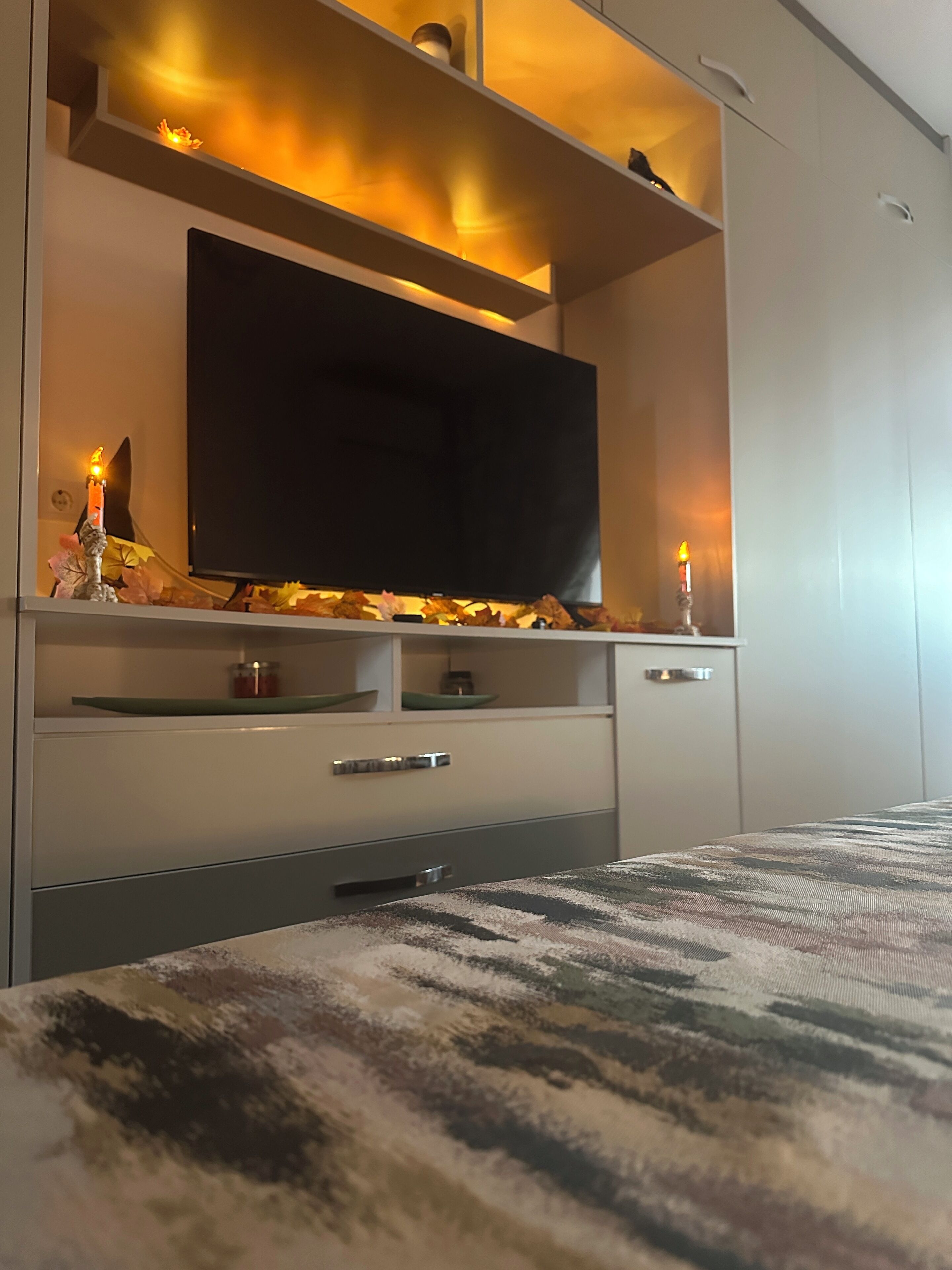 Quarto moderno com televisão de tela plana na parede, cama com colcha estampada e mesa de cabeceira com uma vela.