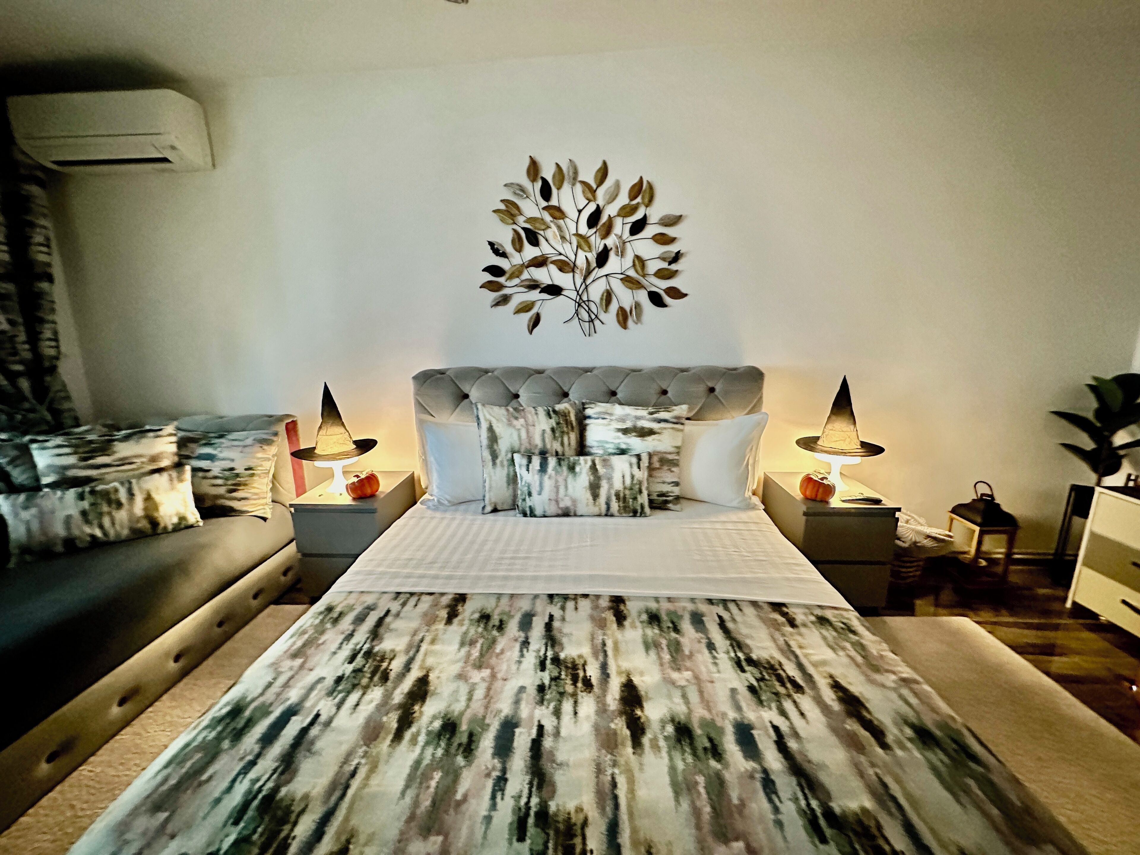 A bedroom with a tufted headboard, a large abstract-patterned bedspread, and two bedside lamps.