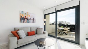 Una sala de estar moderna con un sofá gris, una mesita redonda y un amplio ventanal corredizo que da a una terraza exterior.