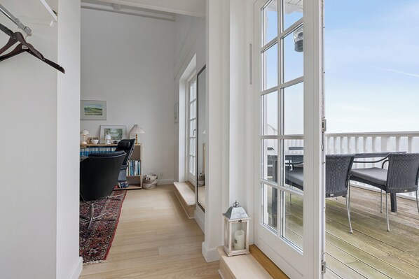 A spacious corridor with a wooden floor, a black chair, a small table, and a window leading to an outdoor deck with wicker furniture. - Appartment by the sea and family friendly beach (Bagenkop)