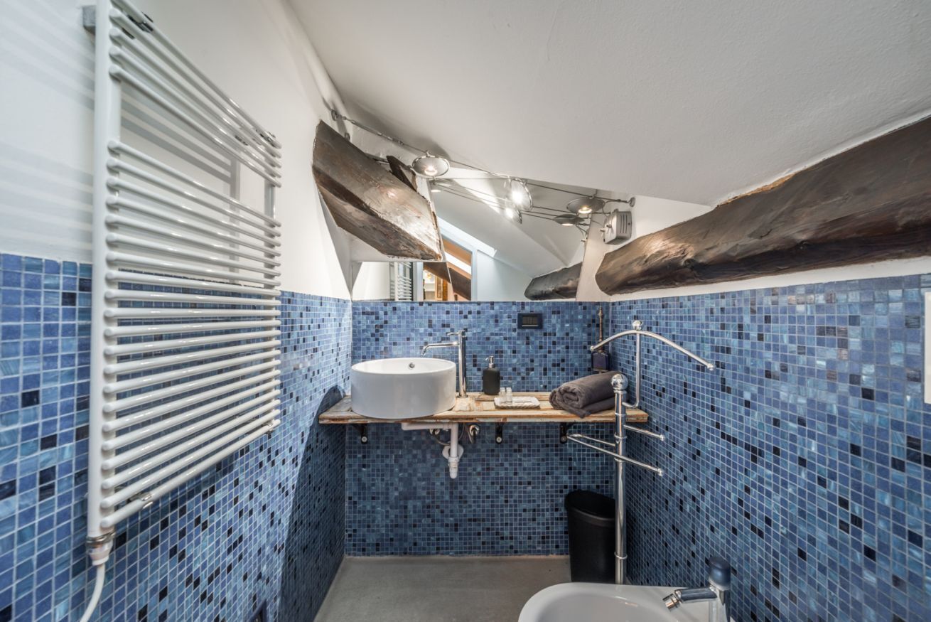 Un bany amb una paret de mosaic blau, un lavabo blanc, una estant de fusta, un penjador de tovalloles i una zona de dutxa.