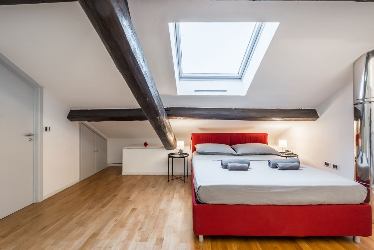 A bedroom with a skylight, a red bed, wooden flooring, and a white bedside table.
