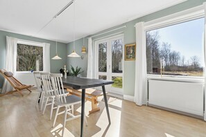 A dining area with a table and chairs, a hanging light fixture, and a view through large windows. - Spacious holiday home on a large natural plot (Nexø)