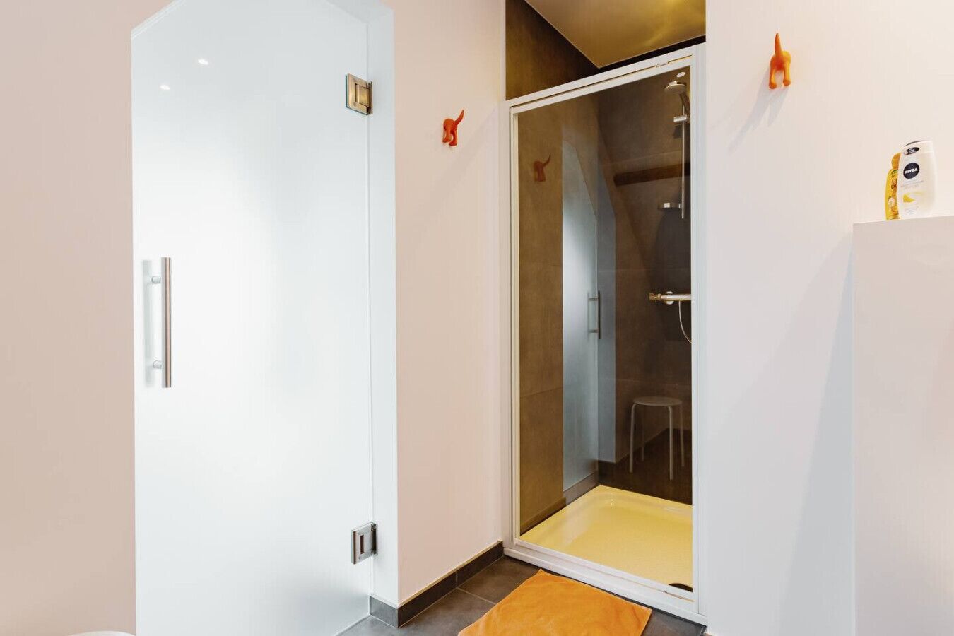 Een moderne badkamer met een douche met glazen wand, een witte deur en een klein krukje.
