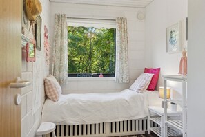 Et lille, hyggeligt soveværelse med en enkelt seng, et vindue med gardiner, et natbord med en lampe og en vægmonteret hylde.