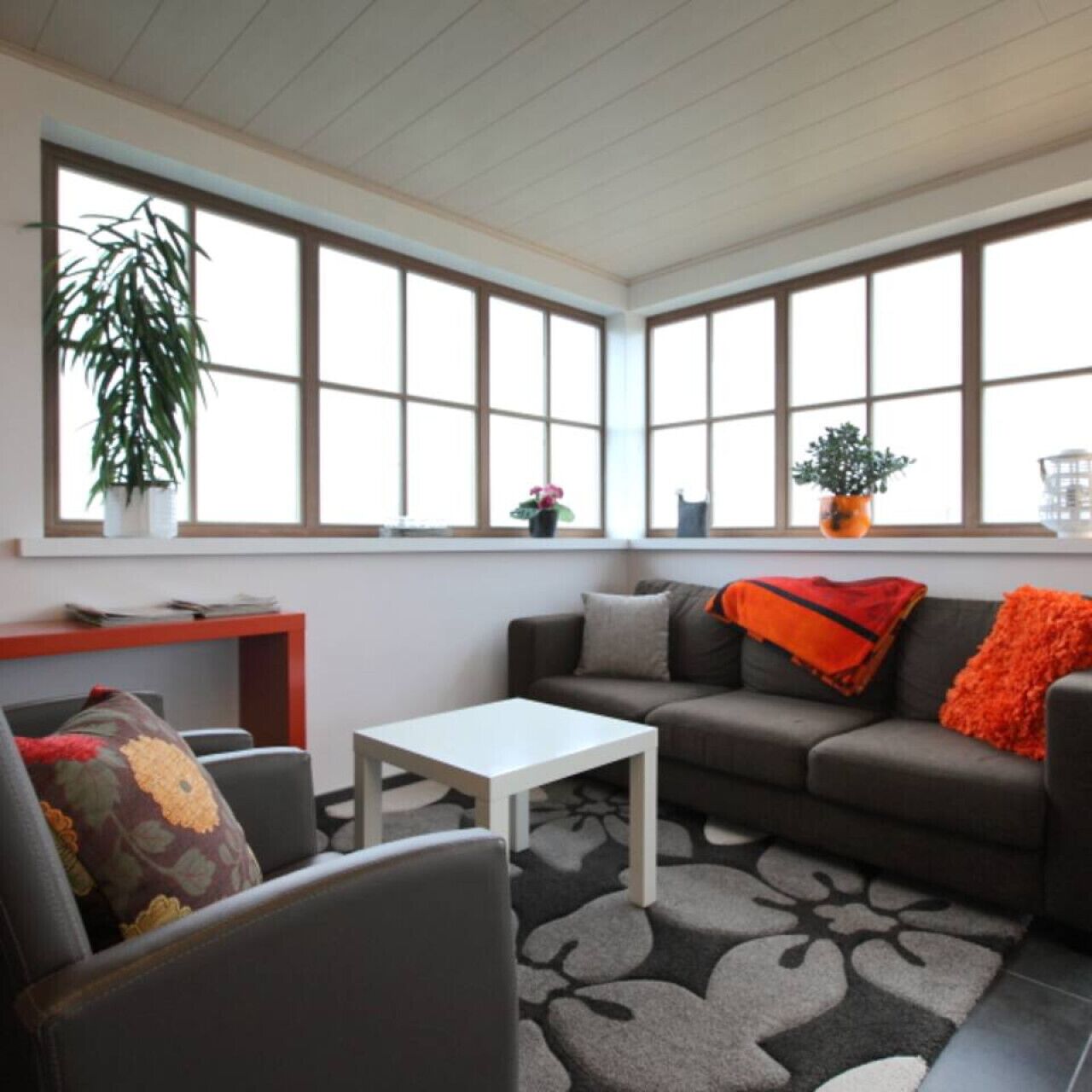 Un salon avec un canapé, une table basse et un fauteuil. De grandes fenêtres sont équipées de stores, une plante est posée sur le rebord de la fenêtre, et une petite table est ornée de divers objets.