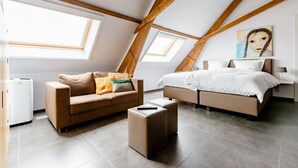 A modern loft bedroom with a sloped ceiling, large windows, a bed, a sofa, and a small table. - Xtra space Deluxe room 1st floor (Ieper)