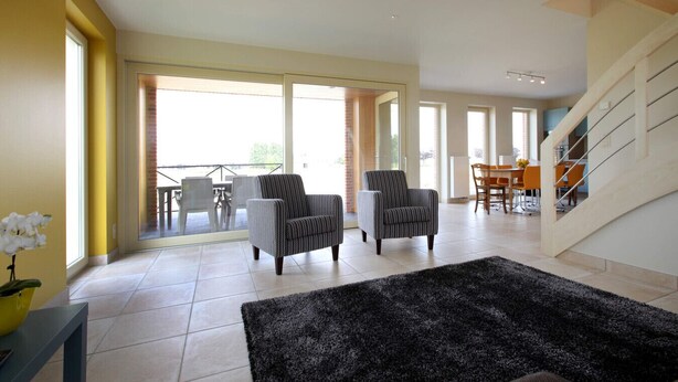 Een ruime woonkamer met twee fauteuils, een zwart vloerkleed en een eethoek met een tafel en stoelen.