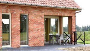 Een bakstenen gebouw met grote glazen deuren, een terras met een tafel en stoelen, en uitzicht op het omliggende landschap.