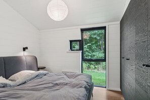 A modern bedroom with a bed, a bedside table, a wall-mounted lamp, a window with a view of greenery, and a wardrobe with a wood-like pattern. - Bright holiday home near Ordrup (Fårevejle)