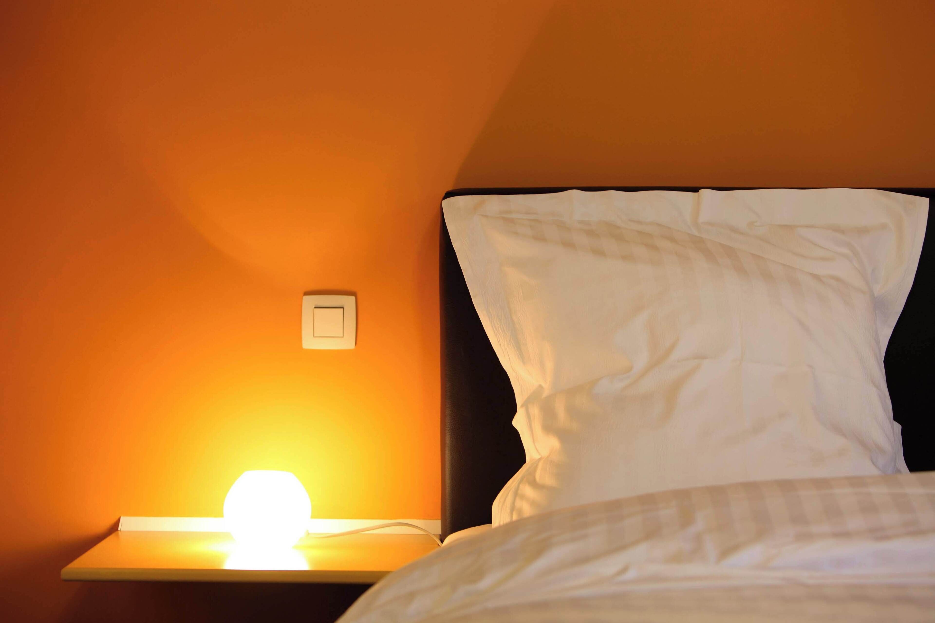 Ein Bett mit weißer Bettwäsche, ein Nachttisch mit einer Lampe und ein Lichtschalter an der Wand.