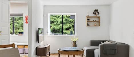 A room with a wooden floor, a round coffee table, a grey sofa, a TV on a stand, and a window with a view of greenery.