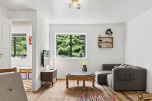 A room with a wooden floor, a round coffee table, a grey sofa, a TV on a stand, and a window with a view of greenery. - Modernised 70s cottage in forest near beach (Nykøbing Sjælland)