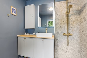 Une salle de bain avec un lavabo blanc, un miroir, une douche dotée d’un pommeau doré et un meuble avec des poignées dorées.