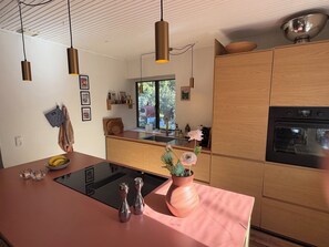 A modern kitchen with a central island, stainless steel appliances, wooden cabinets, pendant lighting, and a view of the outdoors. - Modernised 70s cottage in forest near beach (Nykøbing Sjælland)