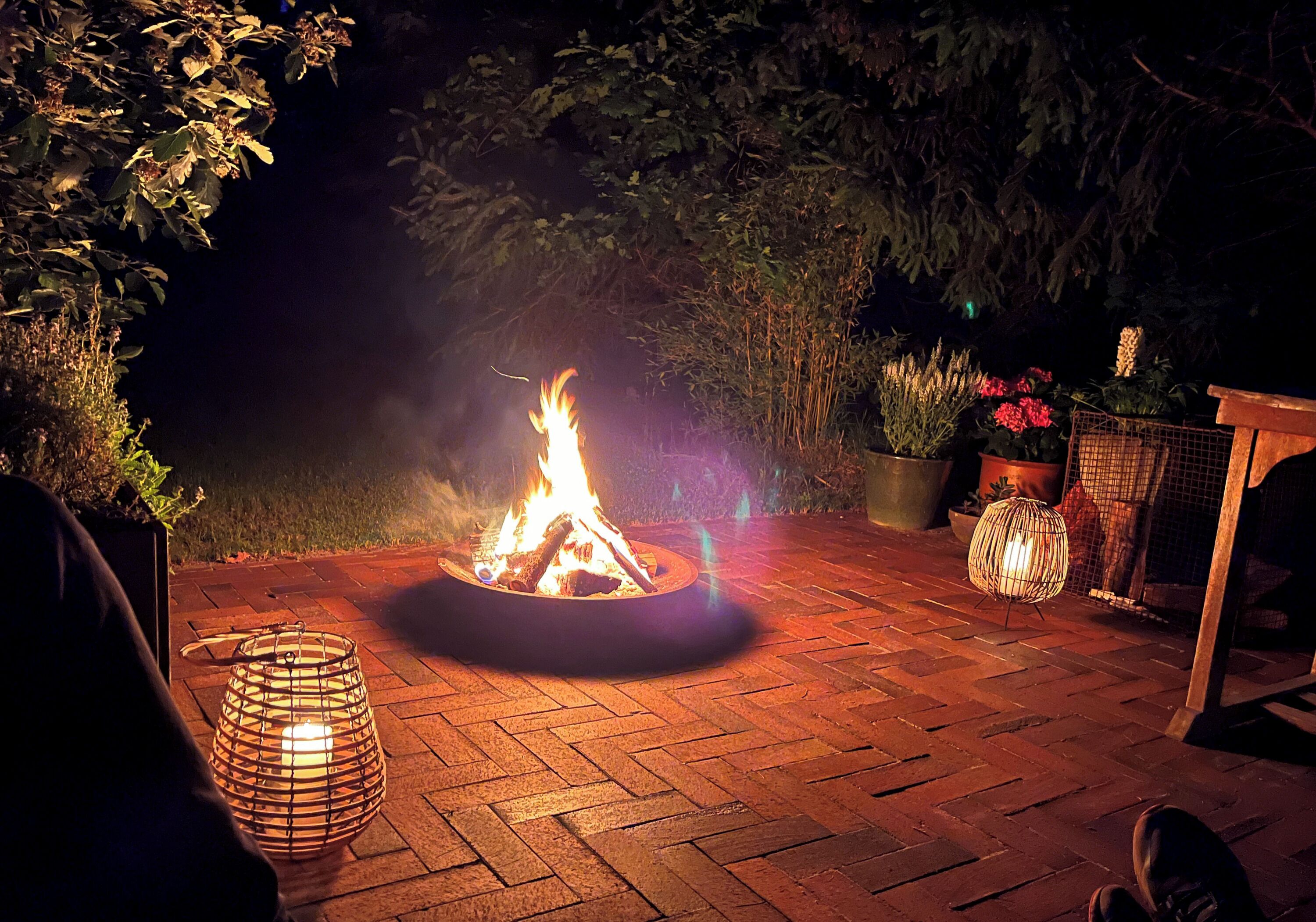 A fire pit in a brick patio area surrounded by potted plants and a lantern.