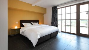 A modern bedroom with a large bed, a nightstand, a wall-mounted light, and a sliding glass door leading to an outdoor area. - Ground floor Standard room - Musterd (Ieper)