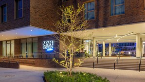 A hotel entrance with a modern brick facade, a neon sign, and a landscaped area with a tree.