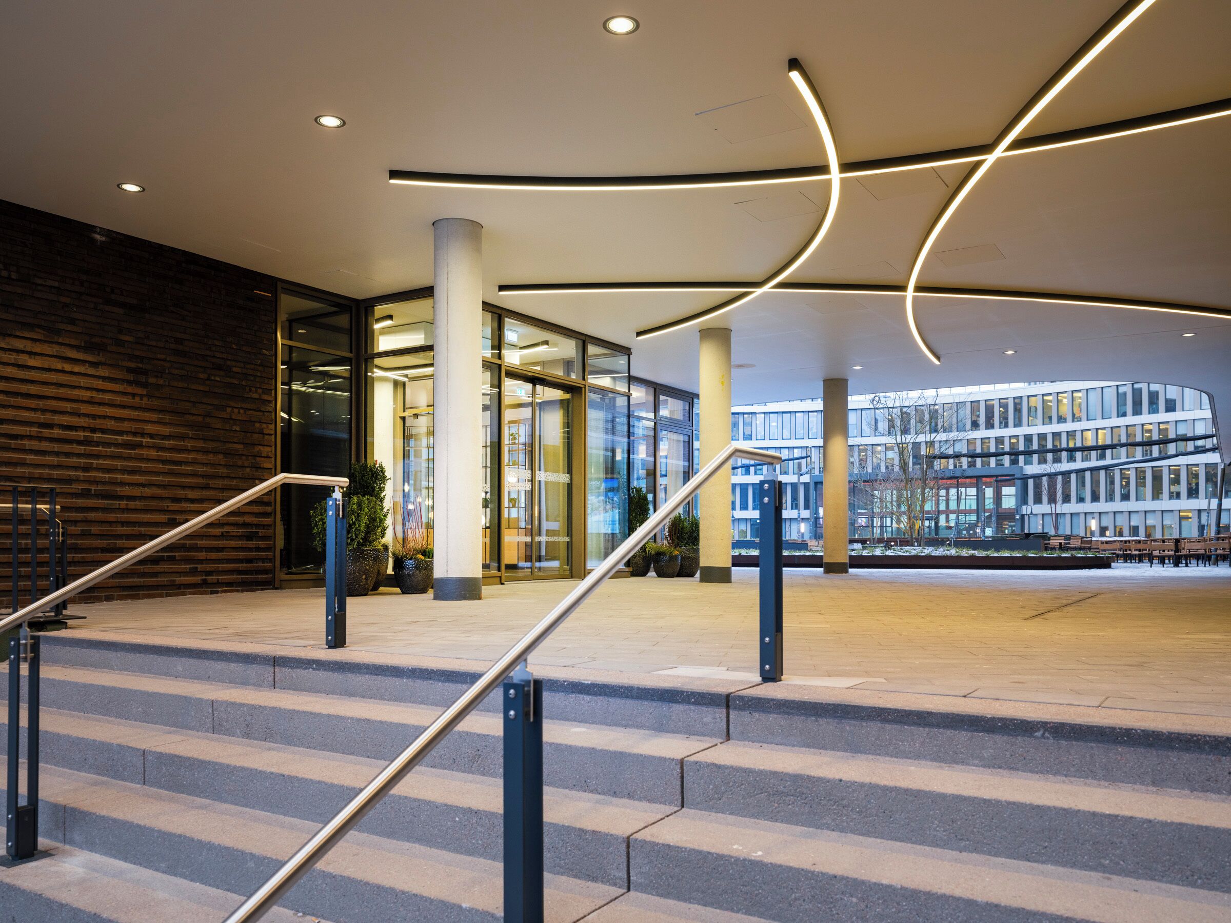 Ein moderner Gebäudeeingang mit Treppe, Glasüren und Blick auf ein anderes Gebäude.