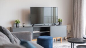 Ett modernt vardagsrum med en platt-TV placerad på ett grått skåp, två krukväxter, en blå pall och en liten träbänk.