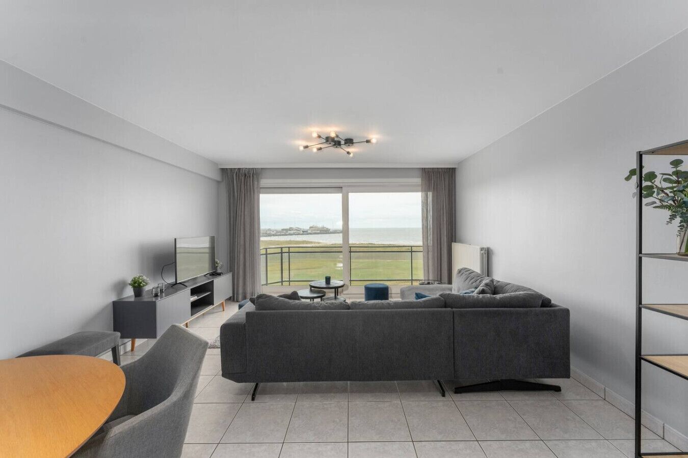 Ett modernt vardagsrum med en stor grå hörnsoffa, ett träbord, en tv och en balkong med utsikt över havet.