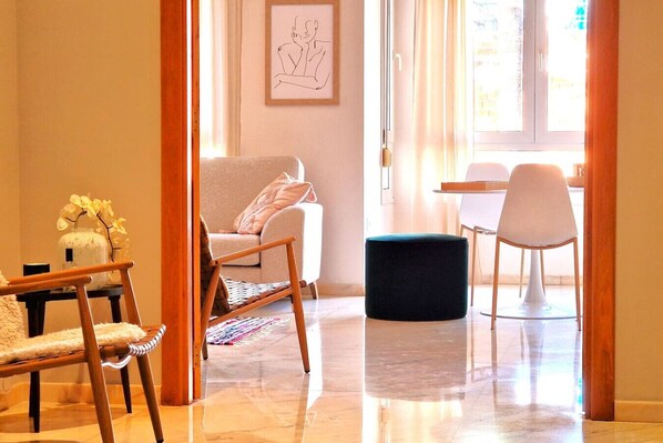 A modern living room with a sofa, a wooden chair, a black ottoman, and a dining area with a table and chairs.