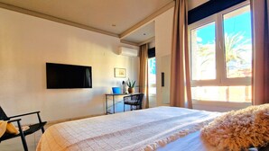 Ein Hotelzimmer mit einem großen Bett, einem Schreibtisch mit einem Laptop, einem Sessel, einem Flachbildfernseher und einem großen Fenster mit Blick auf Palmen.