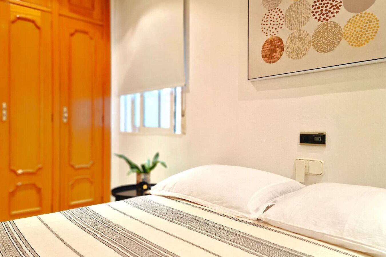 Habitación de hotel con cama, mesita de noche, planta en maceta y termostato en la pared.