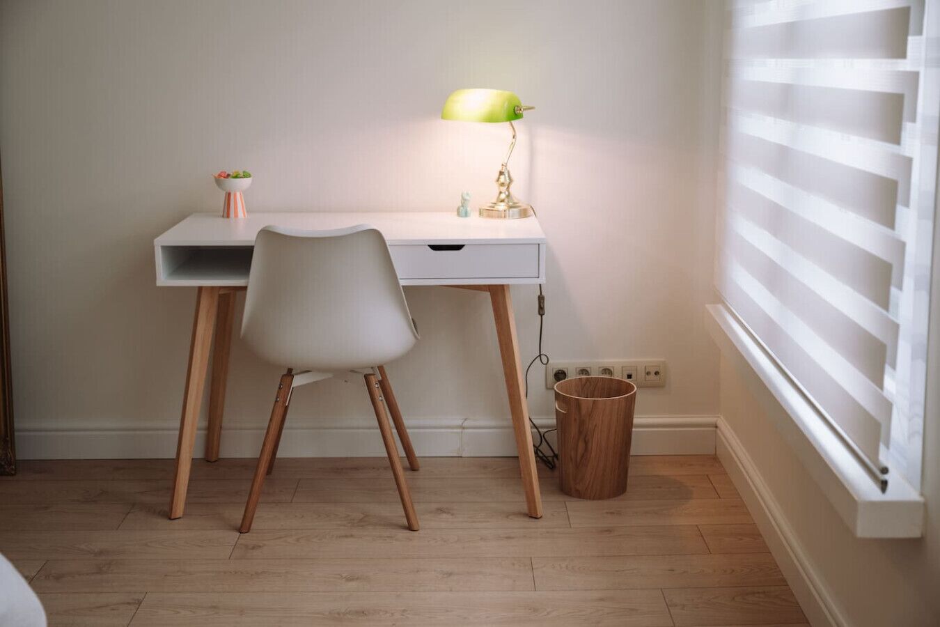 Ein schlicht gestalteter Arbeitsplatz mit einem weißen Schreibtisch, einem weißen Stuhl, einer grünen Schreibtischlampe und einer kleinen Pflanze.