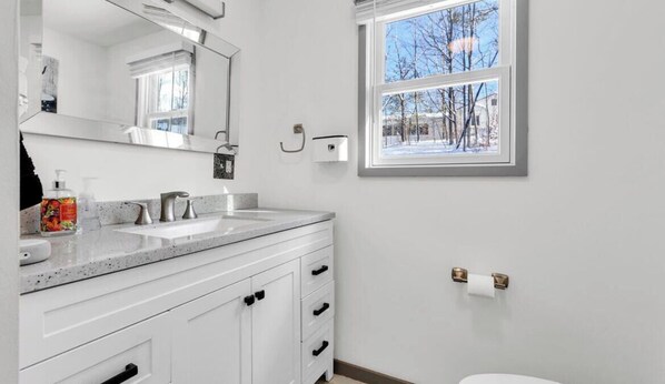 Un bagno con mobili bianchi, un doppio lavabo, uno specchio grande, una finestra vista alberi e un water con dispenser per asciugamani.