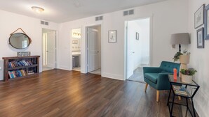 Un amplio salón con suelo de madera, un sillón verde azulado, una mesita auxiliar con una lámpara y una estantería.