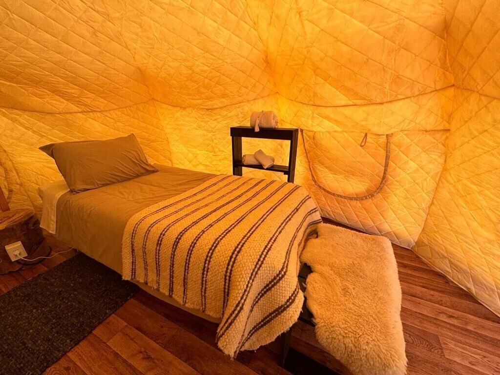 A cozy tent-style room with a bed, a small table with a lamp, and a bench.
