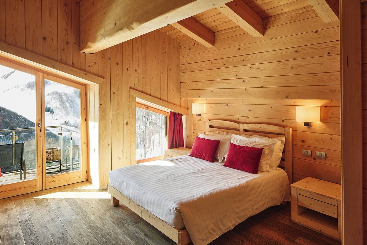 A wooden cabin bedroom with a large bed, a nightstand, a window with a view of a snowy landscape, and a balcony.