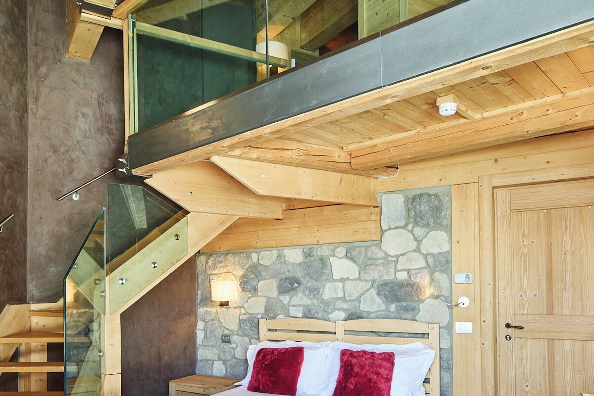A bedroom with a bed, a stone wall, a wooden staircase, and a wooden ceiling.