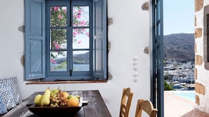 A room with a wooden table, a bowl of fruit, a window with blue shutters, and a view of a town and hills.