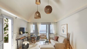 Een moderne woonkamer met een bank, voetenbankje, eettafel, stoelen en een televisie. Er zijn rieten hanglampen, een plant en uitzicht op de omgeving.