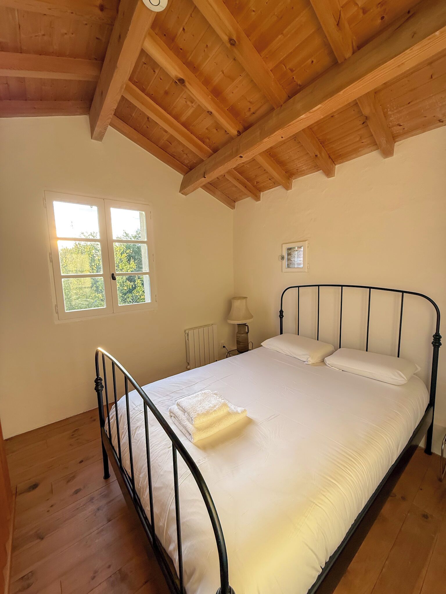 A bedroom with a wooden ceiling, a bed with white bedding, a nightstand, a window, and a radiator.