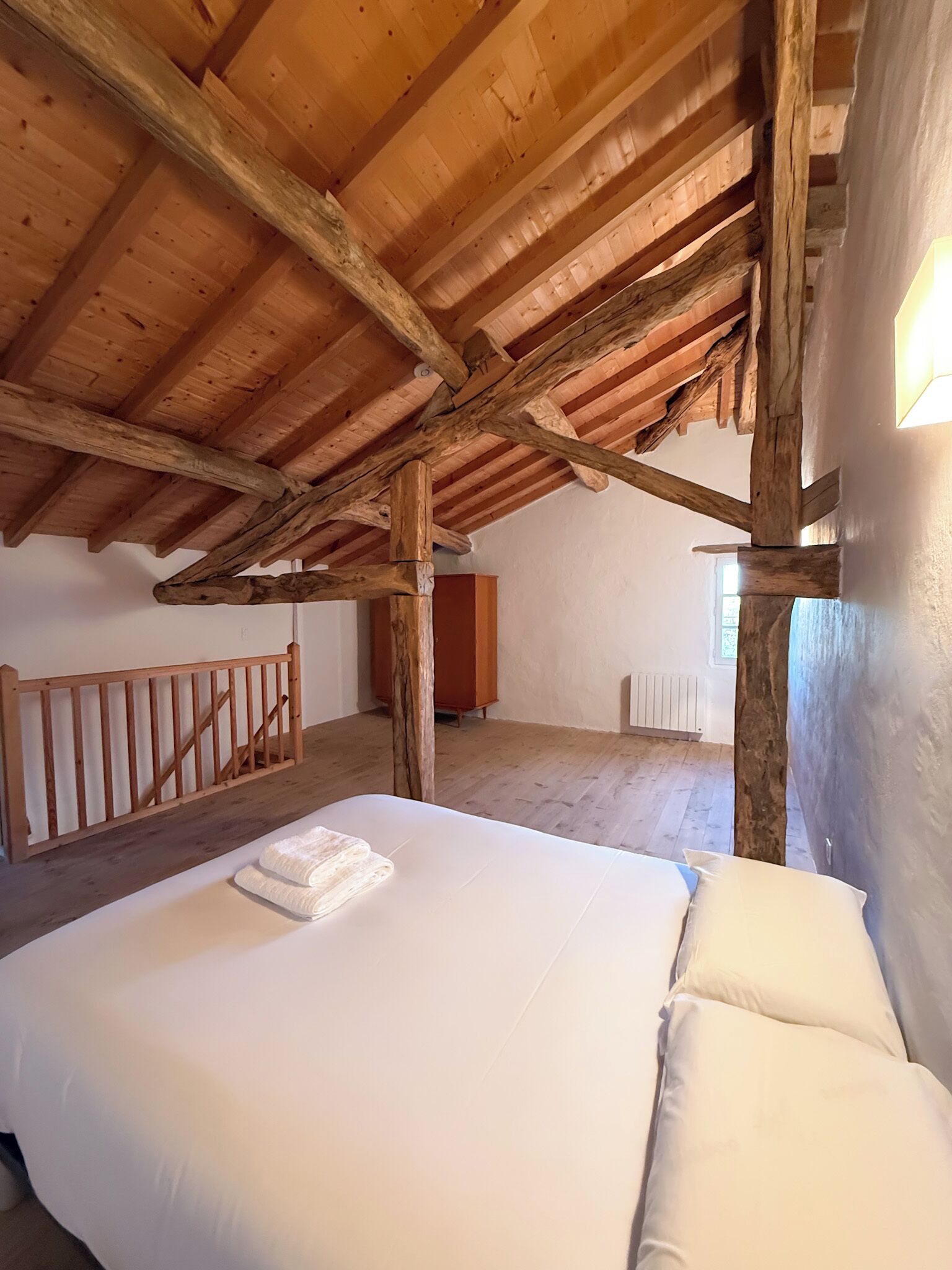 Une chambre avec un plafond en bois, un lit recouvert de draps blancs, une commode en bois et un radiateur.